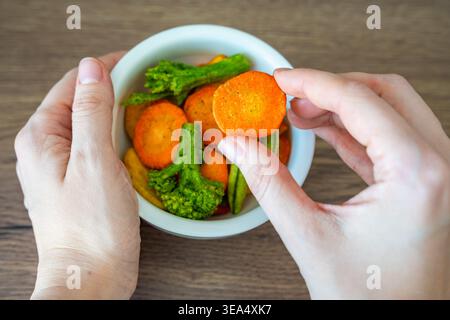 Hand taking carrot vegetable chip from bowl on wooden table. Healthy snack choice and clean eating lifestyle concept. Stock Photo