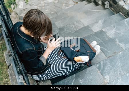 woman sitting on steps with backpack and baguette using smartphone Stock Photo