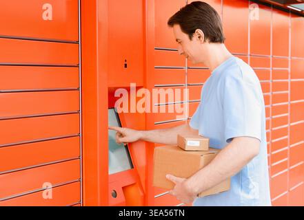 Man with cardboard boxes uses the touch screen of an automated orange parcel locker to pick up or drop off packages. Convenient e-commerce and self-se Stock Photo