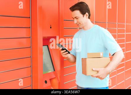 Smiling man using a smartphone at an automated orange parcel locker to pick up or send a package. Holding cardboard boxes, using convenient self-servi Stock Photo