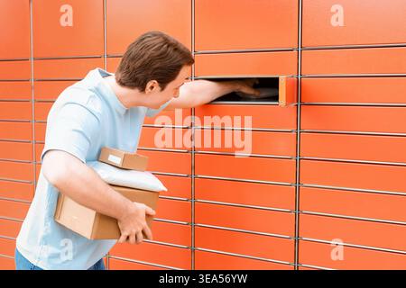 Young man picking up or dropping off packages at an automated orange parcel locker. He holds cardboard boxes, using convenient self-service delivery t Stock Photo