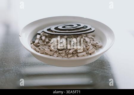 Close-up of spiral mosquito coil resting on fragmented pieces in white ceramic bowl, monochrome Stock Photo