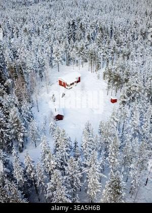 Aerial view of a red wooden cabin surrounded by snow-covered pine trees in a dense winter forest in Lapland, Finland. Stock Photo