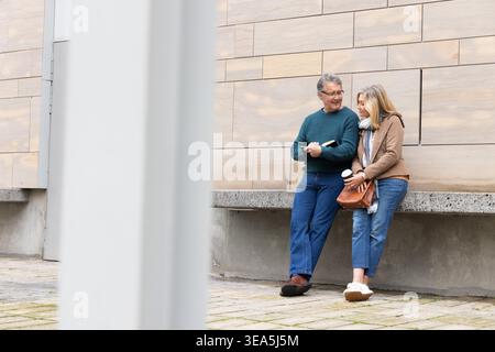 Senior couple leaning against concrete ledge in plaza holding book and coffee cup, copy space. Elderly, duo, urban, notebook, scarf, sidewalk, column Stock Photo