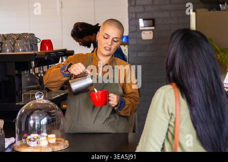Female barista wearing olive apron pouring milk from metal pitcher into red mug at counter. Cafe, espresso, cupcakes, cloche, brick, warmth, bustle Stock Photo