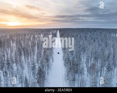 Aerial view of a car driving on a snow-covered road through a dense pine forest during sunset Lapland, Finland. Stock Photo
