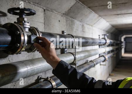 A worker in a high-visibility vest adjusts a digital device attached to a large diameter pipe system within a utility tunnel Stock Photo