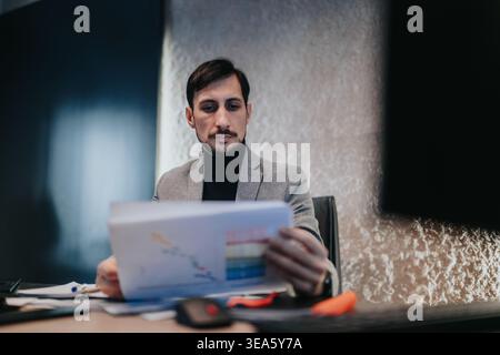 Businessman in blazer reviewing documents at desk in modern office with focused expression Stock Photo