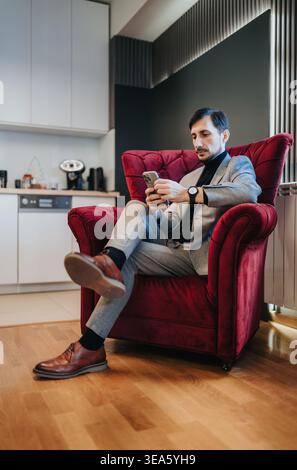 Businessman in gray suit sitting in red armchair using smartphone in modern office kitchen Stock Photo