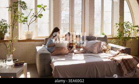 Wide shot of mother and two children using laptop and tablet on sofa in bright living room. Stock Photo