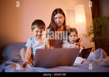 Mother and children using laptop and tablet together on bed in evening light. Stock Photo
