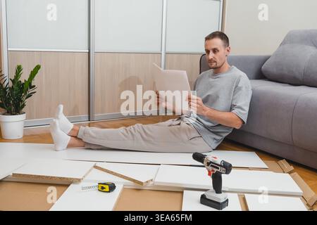 man sits on the floor reading furniture assembly instructions with tools and flat-pack parts scattered around him in a bright, modern living room Stock Photo