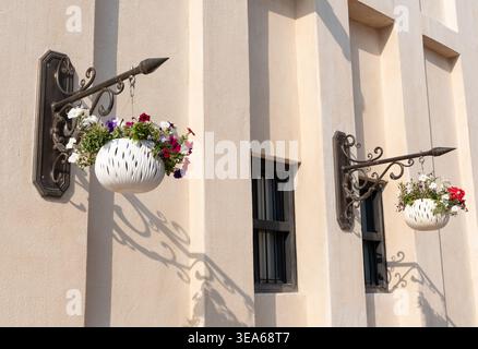 Elegant white floral planters hang from ornate wrought iron brackets on a cream-colored building. Soft afternoon sunlight creates striking shadows Stock Photo