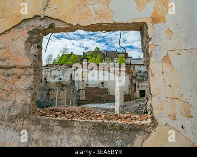 View through a broken wall window shows abandoned building with greenery on top against blue sky and clouds during daytime Stock Photo