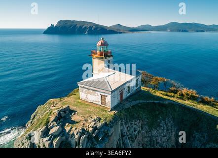 Aerial view captures weathered lighthouse building on a steep coastal cliff by a vast blue ocean beneath a bright sunny sky Stock Photo
