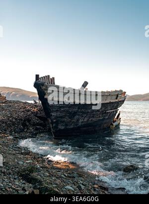 Shipwrecked wooden vessel slowly breaking apart on a stone beach at golden hour near hills Stock Photo