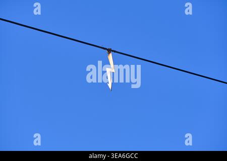 Glowing neon lightning bolt symbol hanging from wire against solid blue twilight sky background Stock Photo