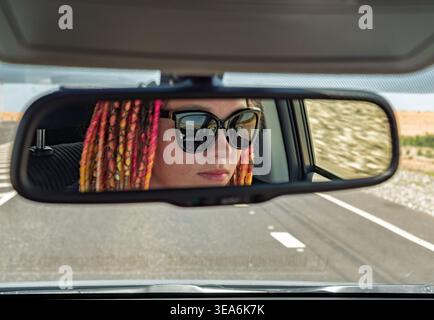 Young woman with bright dreadlocks and sunglasses driving a car reflected in the rearview mirror on a sunny day Stock Photo