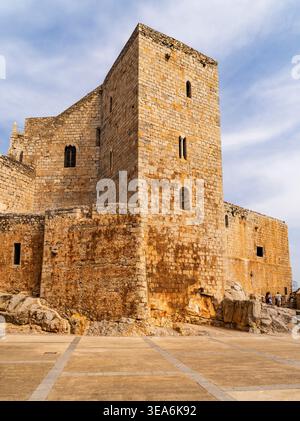 Ancient stone fortress with tall rectangular tower and weathered walls under blue sky, seen from low angle on rocky base with open plaza foreground Stock Photo