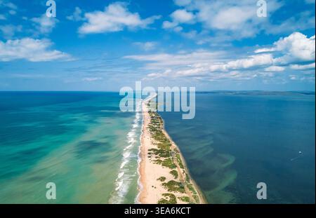 Aerial view of a narrow sandy spit bordered by a wavy turquoise ocean and a calm deep blue lagoon under a bright sky. Stock Photo