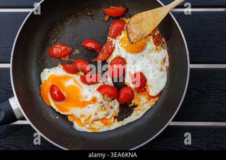 Overhead view of fried eggs cooked with cherry tomatoes in a frying pan on a black table with a wooden spatula. Simple home cooking and breakfast conc Stock Photo