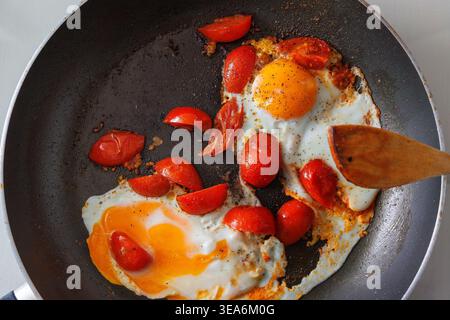 Overhead view of two fried eggs cooked with cherry tomato pieces in a dark frying pan, with a wooden spatula, Simple homemade breakfast. Cooking scene Stock Photo