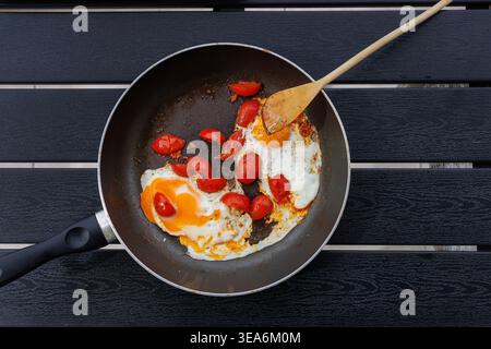 Overhead view of fried eggs cooked with cherry tomatoes in a frying pan on a black table with a wooden spatula. Simple home cooking and breakfast conc Stock Photo