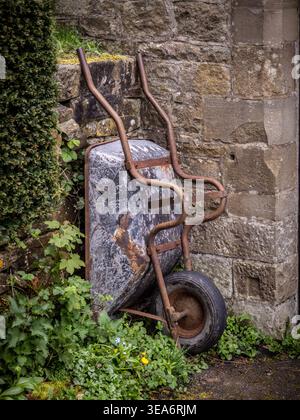 Rusty old wheelbarrow leaning against a stone wall with overgrown plants in a UK garden. Stock Photo
