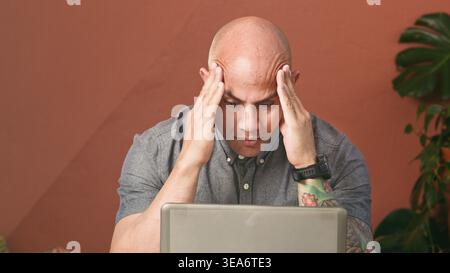 Stressed Remote Worker Concentrating on Laptop With Headache and Tattooed Arm in Home Office Stock Photo
