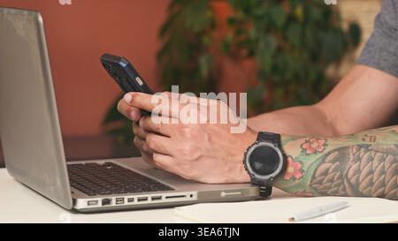 Businessman Using Smartphone and Laptop With Tattooed Arm and Watch in Home Office Stock Photo
