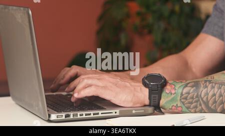 Tattooed Hands Typing on Laptop in Modern Remote Work Workspace With Smartwatch and Notebook Stock Photo