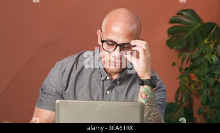 Focused Remote Worker With Laptop and Tattooed Arm Wearing Glasses in Home Office Stock Photo