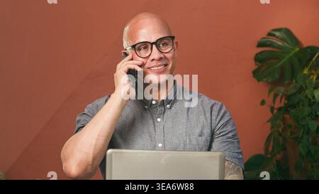 Smiling Businessman Talking on Phone While Working on Laptop at Casual Office Desk Stock Photo