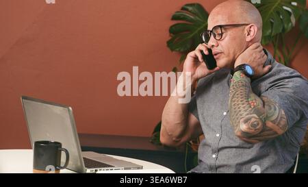 Tattooed Businessman Making Phone Call While Working On Laptop In Modern Home Office Stock Photo