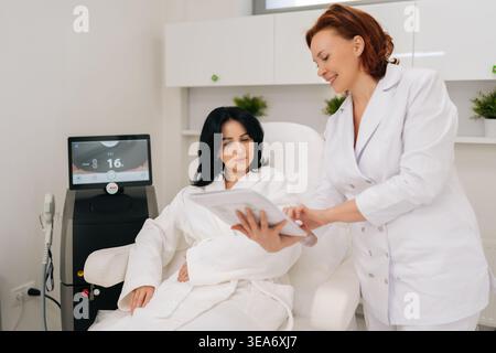 Positive cosmetologist in uniform talking with female client in bathrobe seated in treatment chair, reviewing information on document before cosmetic Stock Photo
