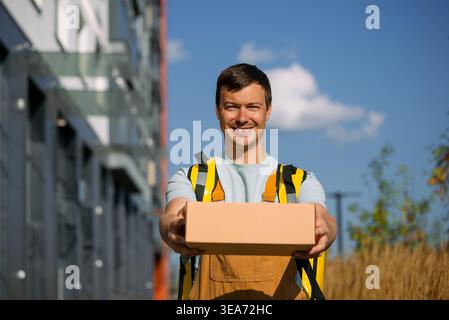 Smiling young courier delivering a package, wearing an insulated delivery backpack and overalls, standing outdoors in front of a modern city building, offering excellent shipping service Stock Photo