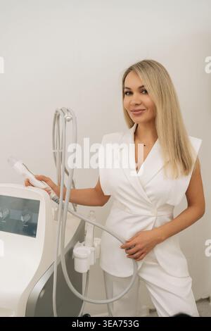 Vertical portrait of blonde esthetician in white coat setting up modern fractional radiofrequency microneedling device, adjusting parameters before Stock Photo