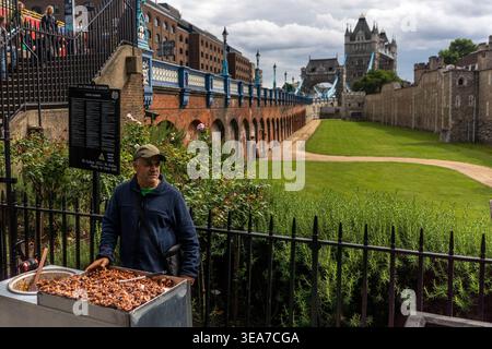 A man stands behind a metal cart filled with roasted nuts, positioned in front of a historic stone wall and a long grassy path leading toward a castle Stock Photo