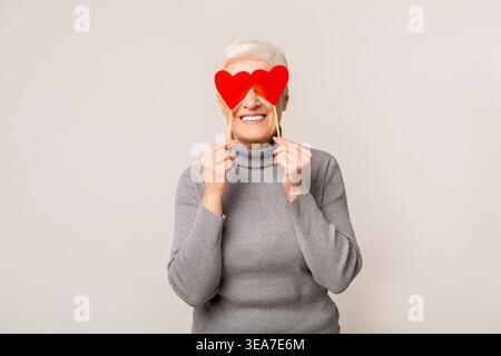 Smiling Elderly Woman Holding Heart Shaped Props for a Fun Celebration in a Light Setting Stock Photo