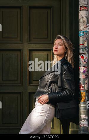 Elegant young woman in a leather jacket and white pants posing against a dark green doorway in warm daylight. Clean composition, stylish accessories, Stock Photo