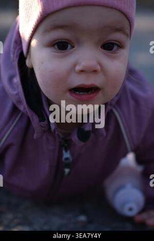 close-up portrait of a curious toddler outdoors in soft winter light, wearing a pink beanie and purple jacket with hood and zipper, leaning forward on Stock Photo