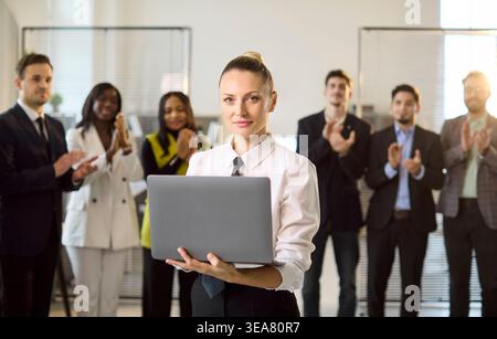 Businesswoman with laptop leads team as colleagues applaud in modern office Stock Photo