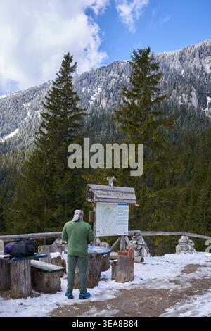 A mature man stands with his back to the camera reading a trail map board at a snowy mountain rest stop with the rocky face of Piatra Craiului rising Stock Photo