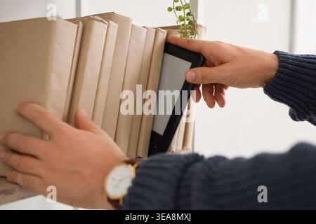 Young man taking e-reader from shelf with books at home, closeup Stock Photo