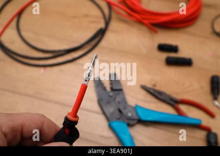 A close-up of a hand holding a red solar cable with a metal contact ready to be inserted into an MC4 connector body. Crimping pliers and spools of red Stock Photo