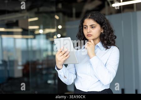 Young professional businesswoman reviewing information on a digital tablet, standing thoughtfully in a modern office environment, contemplating a complex business decision or strategy Stock Photo