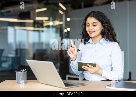 Young indian businesswoman having a virtual discussion or meeting, holding a pen and notebook while gesturing, working on a laptop in a modern corporate office environment Stock Photo
