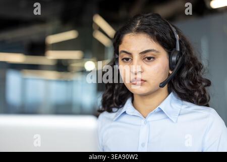 Young indian woman in light blue shirt wearing headset, working concentrated in a modern call center, providing customer support and communication services to clients Stock Photo