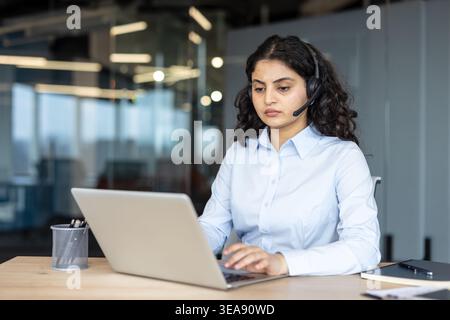 Professional female customer support agent or telemarketer sitting at an office desk, wearing a headset, and actively typing on a laptop, providing service Stock Photo