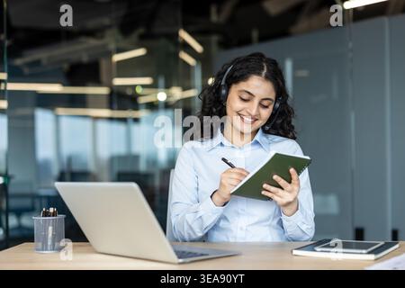 Young businesswoman wearing a headset and smiling, actively writing notes in a notebook while working at a laptop in a modern office, focusing on communication and customer support Stock Photo